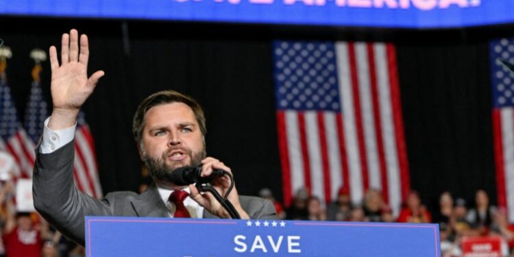 JD Vance raises a hand as he speaks behind a podium that reads,