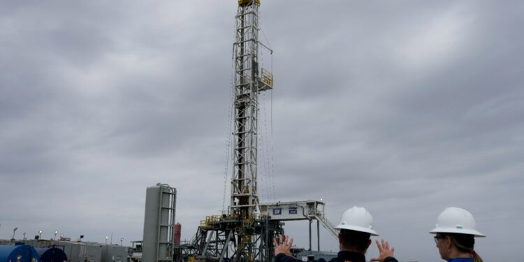 Two people in hard hats survey a drilling rig by Chevron under a grey, cloudy sky,