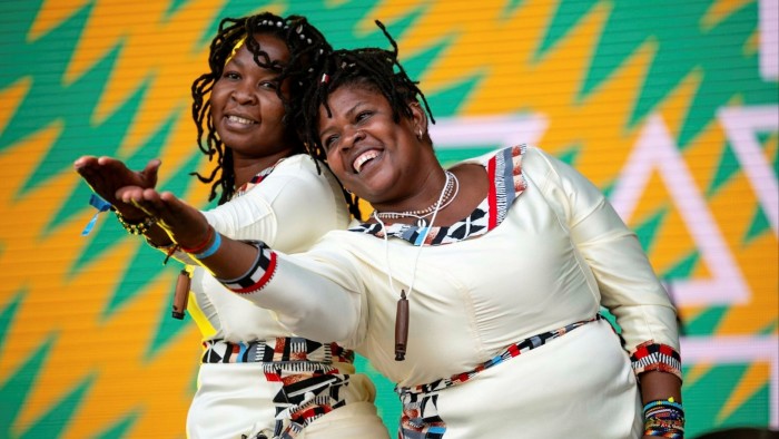 Two women in white dresses smile as they lean against each other and extend an arm forward