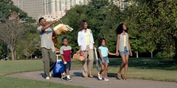 A family of five walking along a path in Piedmont Park, Atlanta, with picnic baskets and a cooler on wheels.