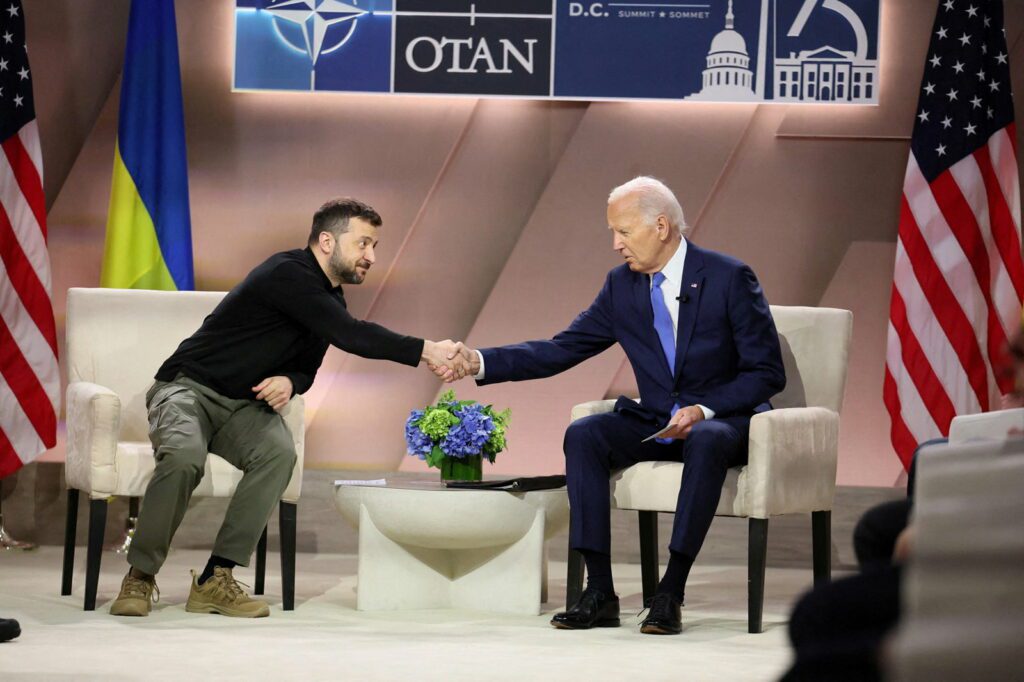 Volodymyr Zelenskyy and Joe Biden, who are seated in chairs, reach across a small table to shake hands.