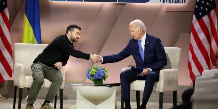 Volodymyr Zelenskyy and Joe Biden, who are seated in chairs, reach across a small table to shake hands.