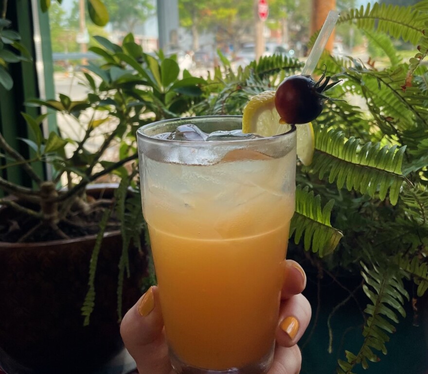 A hand with painted nails holds out a glass of lemonade in front of plants.