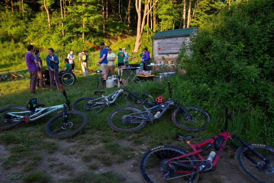 A photo of bikes laying on grass in the foreground with people standing and sitting in wheelchairs in the background. 