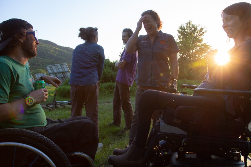 A photo of people outside during golden hour. The grass is green and there are mountains and blue sky in the background. Two people in the foreground are seated in wheelchairs, and three people are standing in the background. Everyone is talking, engaged and smiling. 