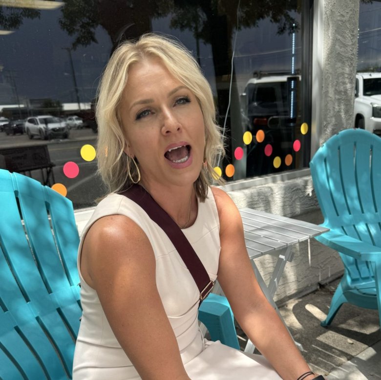 A blonde woman in a white dress, sitting on a turquoise chair, speaks to a person while waiting to testify in Tina Peters' trial.