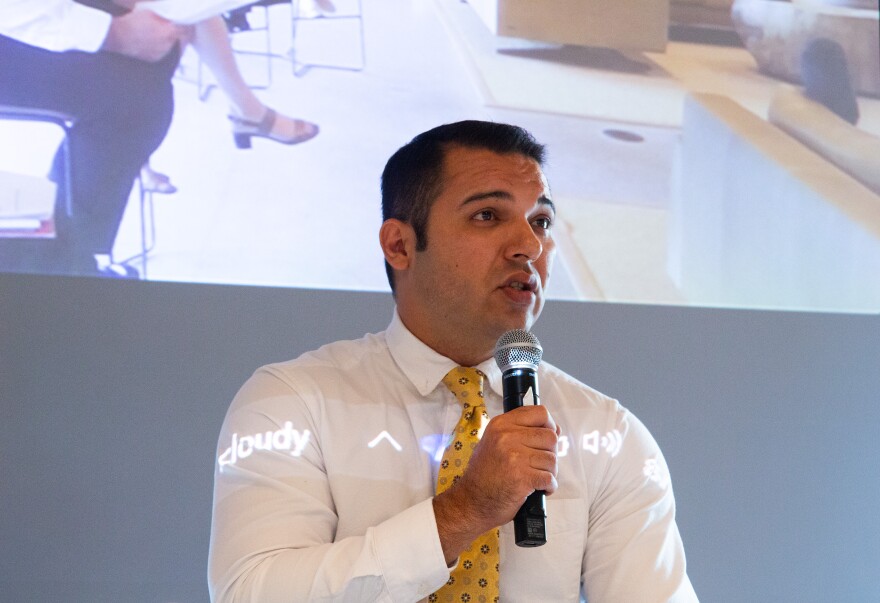 A photo of a man with short black hair wearing a white shirt and yellow patterned tie holds a microphone and speaks into it. behind him is a projected screen with a zoom box showing people sitting in chairs.