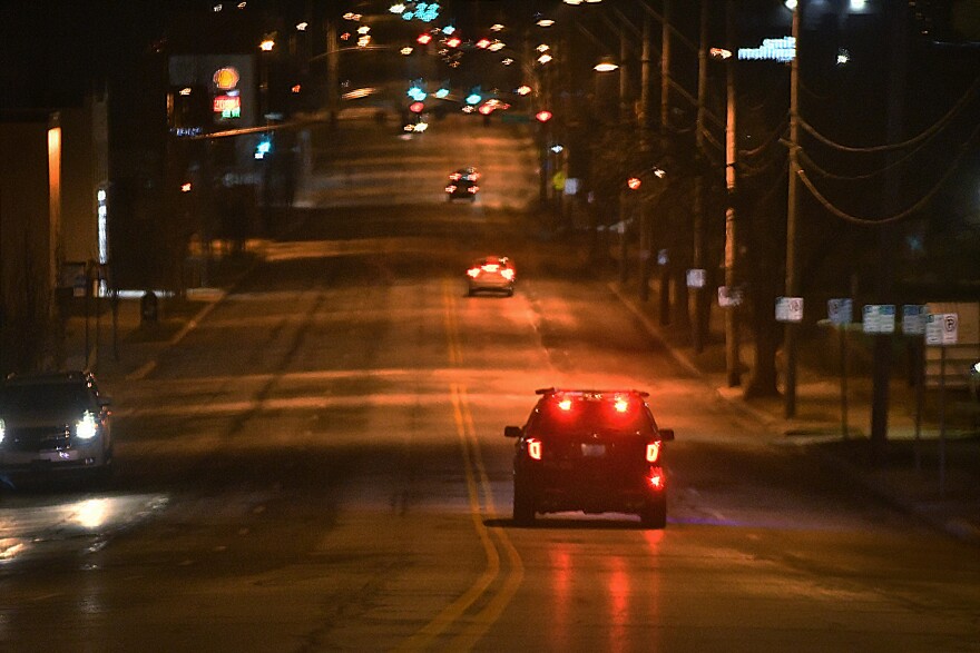 A police cruiser is seen with red lights glowing as it drives down a street at night. There are a few cars on the street, and their tail lights can be seen.