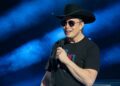 Elon Musk speaks at an event while wearing a cowboy hat, sunglasses, and T-shirt.