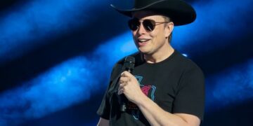 Elon Musk speaks at an event while wearing a cowboy hat, sunglasses, and T-shirt.