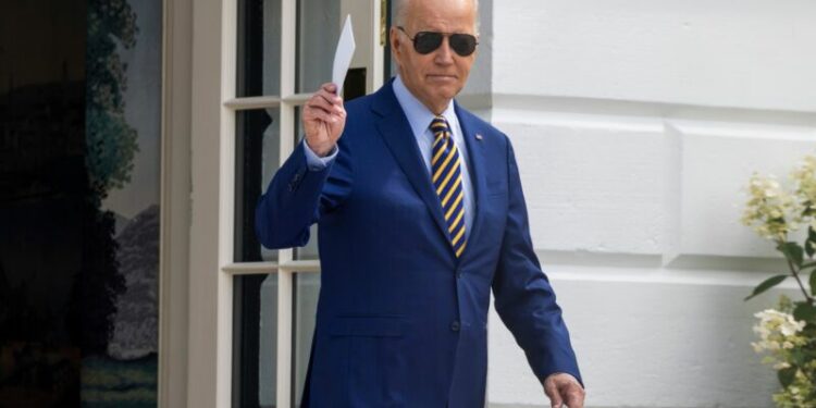 Joe Biden walking outside the White House, wearing sunglasses and holding a stack of index cards in his right hand.