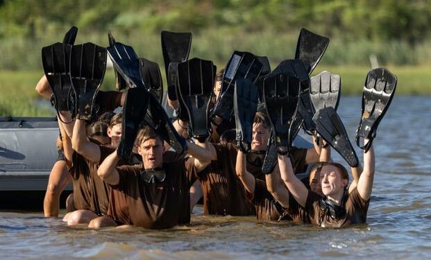 A grouping of students in brown T-shirs wade through chest-high river water holding diving flippers over their heads.