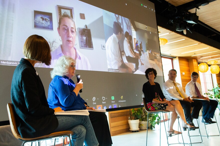 A photo of five people sitting in high chairs in front of a large pull down screen that has two Zoom boxes in it, one of a woman on the left and on the right, a view of the five people in chairs from the side. A person wearing a blue shirt with white hair is speaking into a microphone.