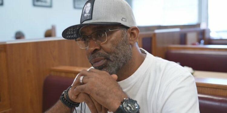 A man wearing glasses, a grey hat and a white T-shirt.