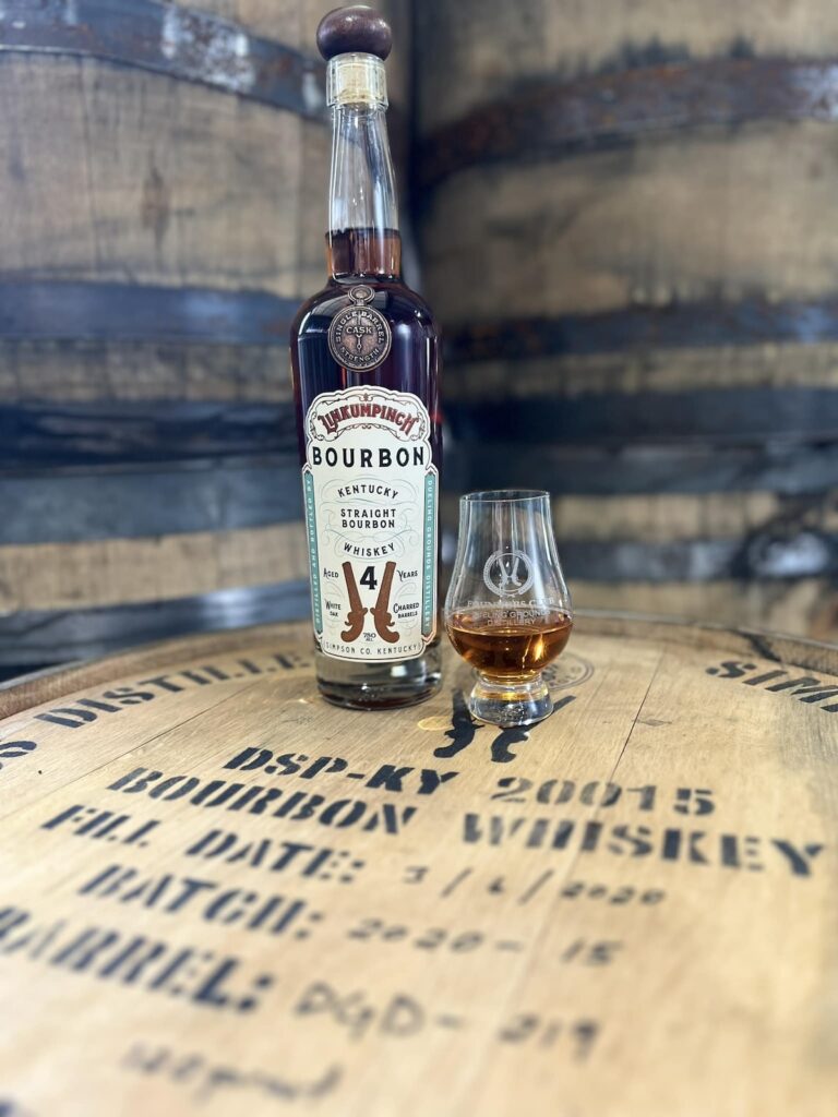 A bottle of 14-year-old Kentucky Straight Bourbon and a glass with a small amount of bourbon sit on a barrel labeled with distillery details. In the background, additional barrels add to the rustic allure.