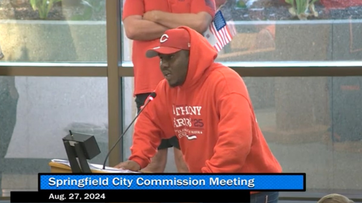 Man in red shirt, ball cap speaking into microphone