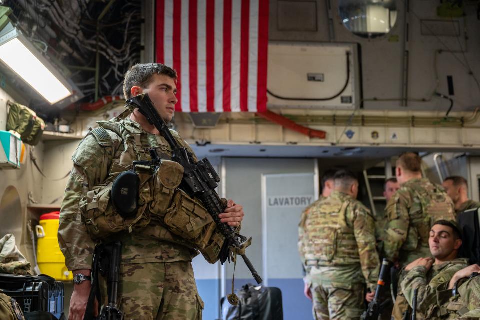 A US Army soldier standing and holding a gun with other soldiers and an American flag in the background.