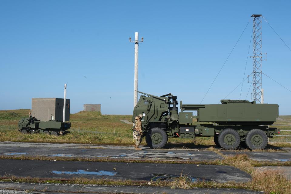 A US Army soldier stands in front of a communications system truck parked on a road near green fields against a blue sky.