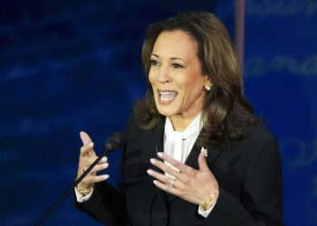 Kamala Harris, wearing a dark suit and white blouse, gestures with her hands as she speaks into a microphone.