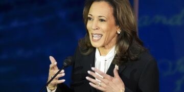 Kamala Harris, wearing a dark suit and white blouse, gestures with her hands as she speaks into a microphone.