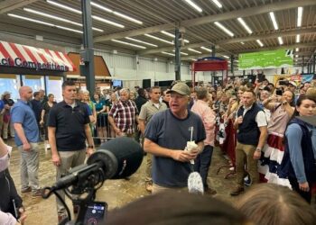 A man stands around a large crowd and media while speaking.