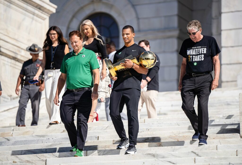 Boston Celtics Coach Joe Mazzulla, a Rhode Island native, walks down the Rhode Island State House steps with the NBA championship trophy along with Celtics co-owner Steve Pagliuca, left, and Rhode Island Governor Daniel J. McKee, right.