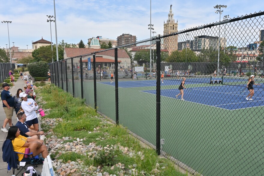 Outdoor photo showing people at left sitting in folding chairs watching several players on many different tennis courts. A city skyline can be seen in the background showing the Kansas City Historic Plaza.