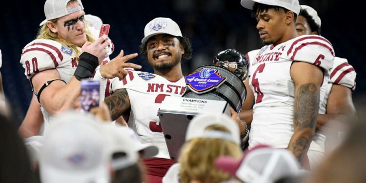 Dec 26, 2022; Detroit, Michigan, USA; New Mexico State University linebacker Trevor Brohard (left) takes a picture of safety Bryce Jackson (center) holding the Quick Lane Bowl trophy on the podium during their post-game celebration after their win over Bowling Green State University in the 2022 Quick Lane Bowl at Ford Field. Mandatory Credit: Lon Horwedel-USA TODAY Sports