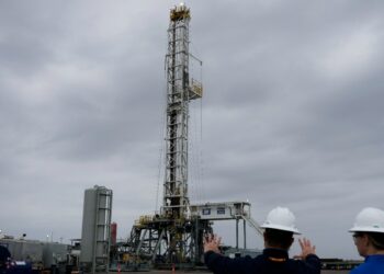 Two people in hard hats survey a drilling rig by Chevron under a grey, cloudy sky,