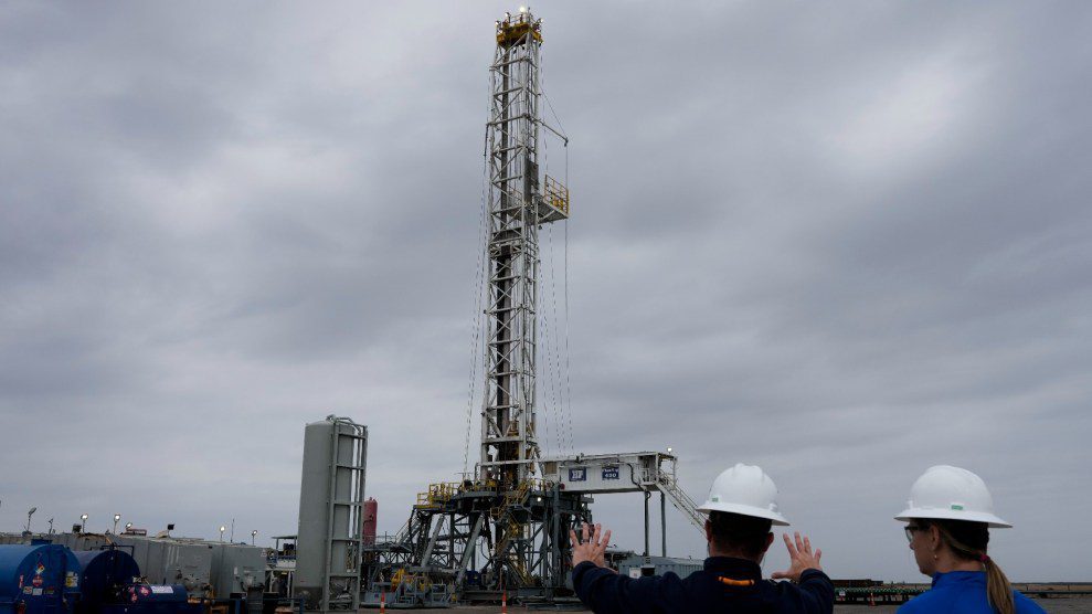 Two people in hard hats survey a drilling rig by Chevron under a grey, cloudy sky,