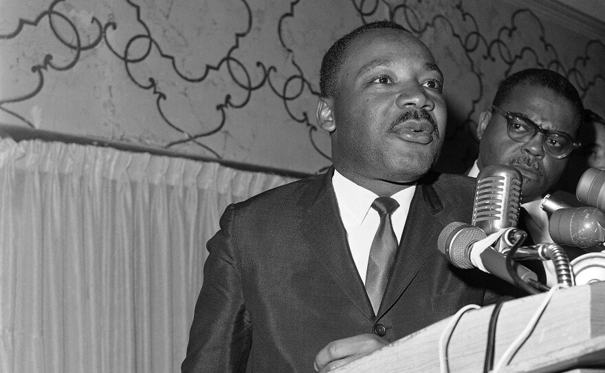 Dr. Martin Luther King Jr., speaks at a podium while Rev. O. M. Hoover looks on.