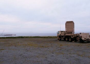 A US Army Q-53 Radar is parked on a sandy shore overlooking the ocean and a grey sky.