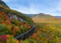 Aerial drone photo of during autumn day of the beautiful red, orange and yellow leaf foliage. Taken in the White Mountains, New Hampshire with train track trestle curving around mountainside.