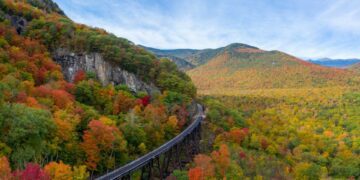 Aerial drone photo of during autumn day of the beautiful red, orange and yellow leaf foliage. Taken in the White Mountains, New Hampshire with train track trestle curving around mountainside.