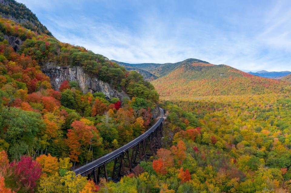 Aerial drone photo of during autumn day of the beautiful red, orange and yellow leaf foliage. Taken in the White Mountains, New Hampshire with train track trestle curving around mountainside.